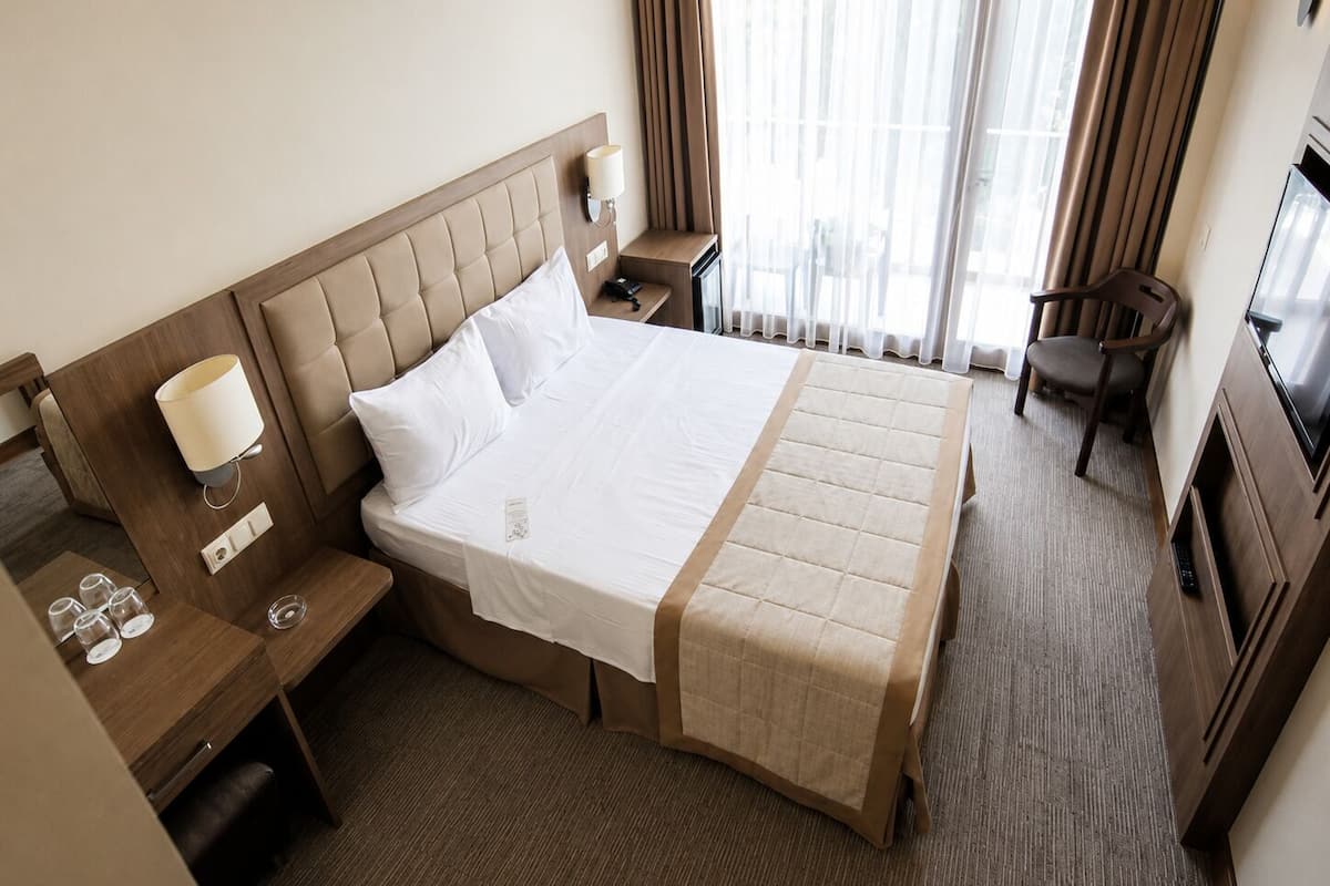 A hotel room with a large bed, bedside tables, a desk, a chair, a TV, and a window with curtains.
