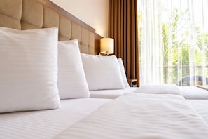 Habitación de hotel con una cama grande, varias almohadas, un cabecero de madera, una lámpara de noche y vistas a árboles por la ventana.
