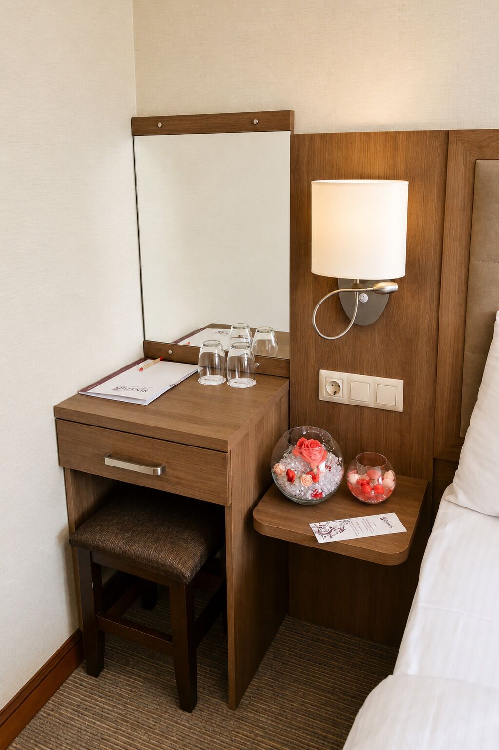 A hotel room with a wooden desk, a bedside table with a lamp, a chair, and a bed.
