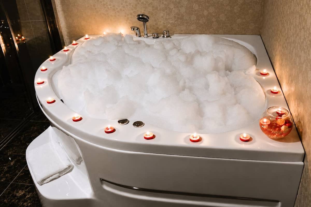 A bathtub filled with bubbles, surrounded by candles, and a glass bowl with candles inside.