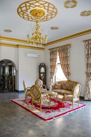 Een kamer met een kroonluchter, een tapijt met een rood-wit patroon, een gouden divan en een houten kast.