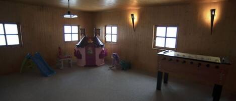 A room with wooden walls, a ceiling fan, a foosball table, a pink playhouse, and windows.