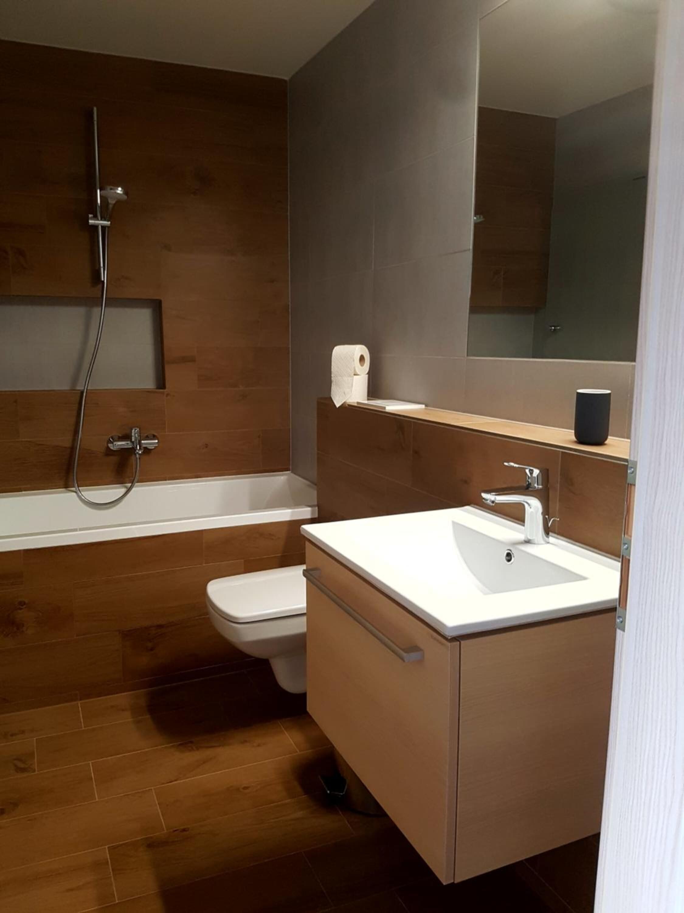 A modern bathroom with a wooden floor, a white toilet, a bathtub, and a sink with a mirror above it.
