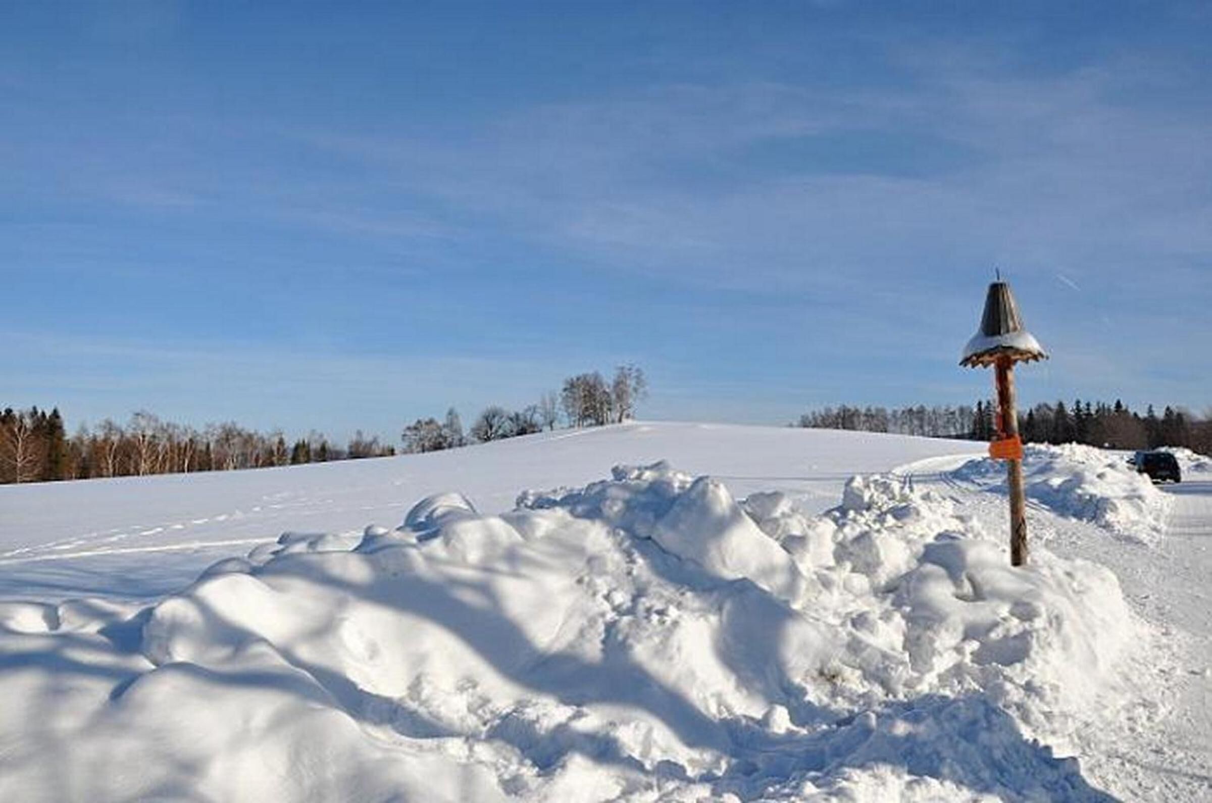 Schneebedeckte Landschaft mit einem hölzernen Pfosten und einer Metallkonstruktion obenauf, vermutlich ein Schild oder eine Wegmarkierung, im Vordergrund.