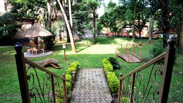 A garden pathway leading to a gazebo with a thatched roof, surrounded by lush greenery and a small bridge.