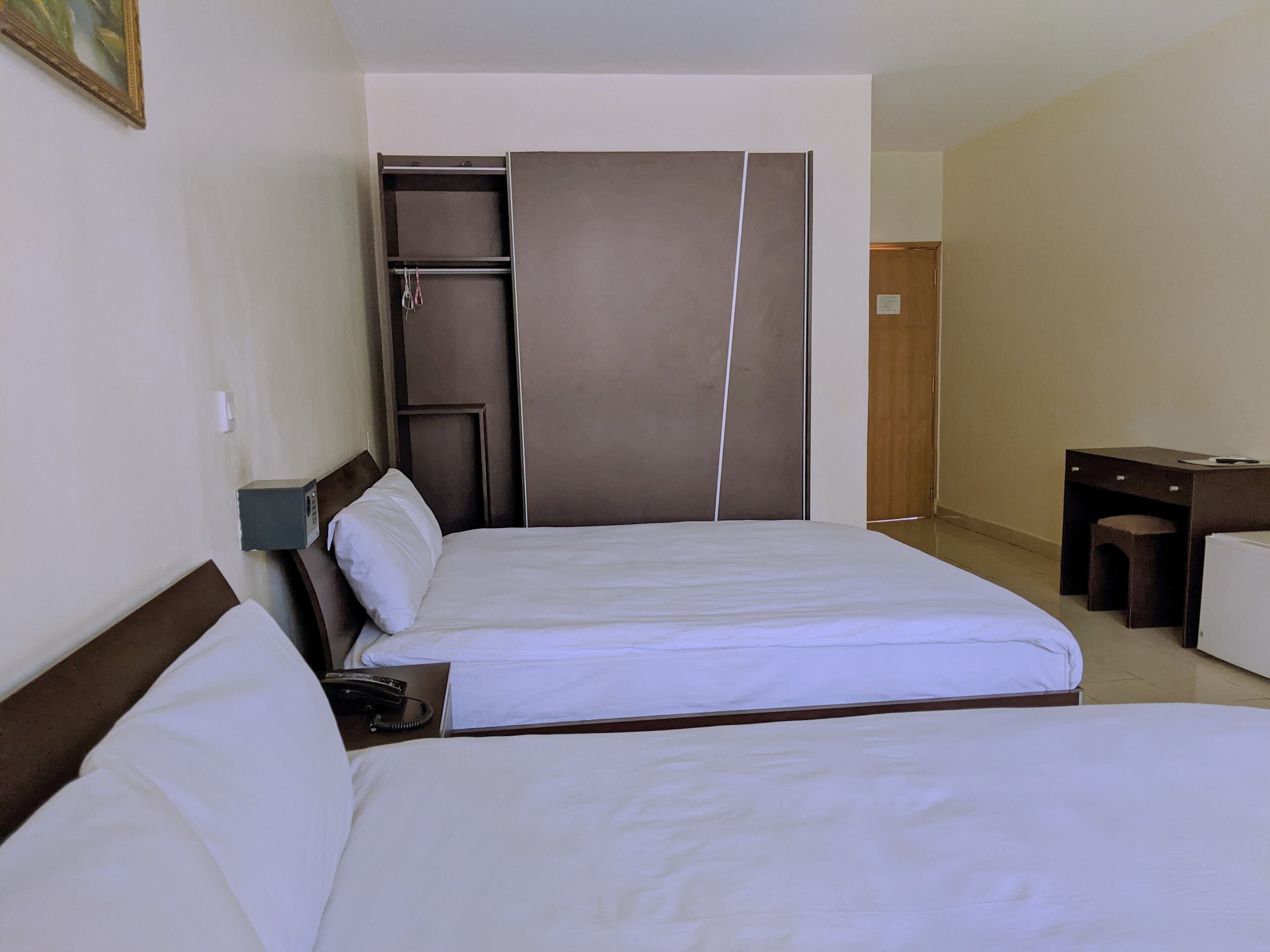 A hotel room with two beds, a desk, a wardrobe, and a small stool.