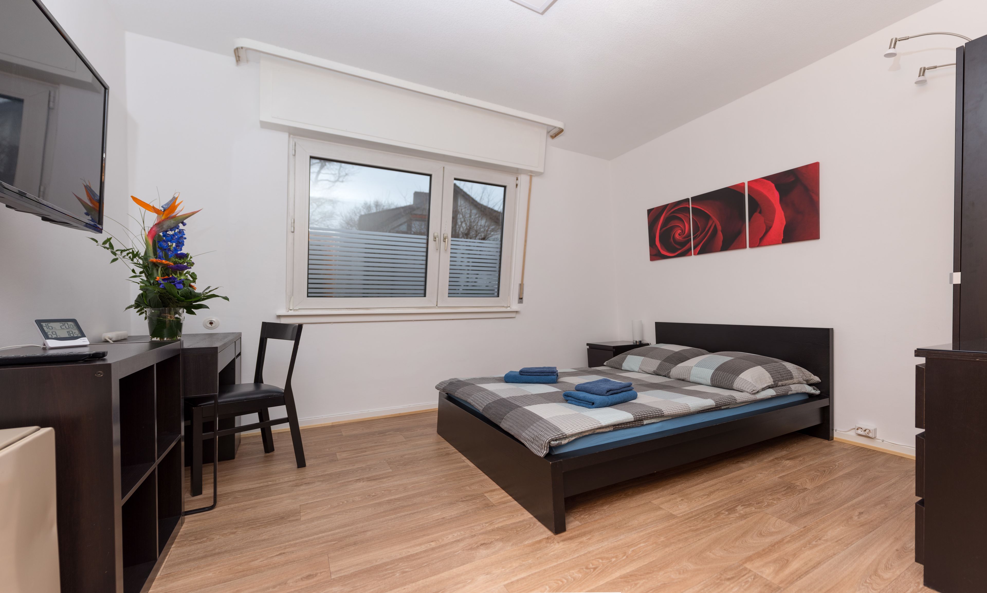 Ein modernes Schlafzimmer mit Bett, Schreibtisch und Stuhl. Es gibt ein Fenster mit Jalousien, ein Wandbild und eine kleine Pflanze auf dem Schreibtisch.