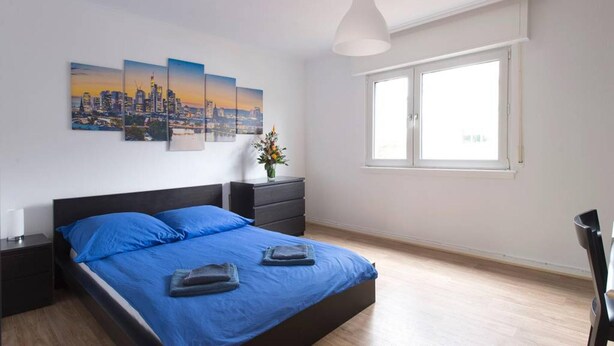 A modern bedroom with a large bed, a cityscape mural, a vase with flowers, and a window with natural light. - Schaefer Apartments GmbH (Muehlheim am Main)