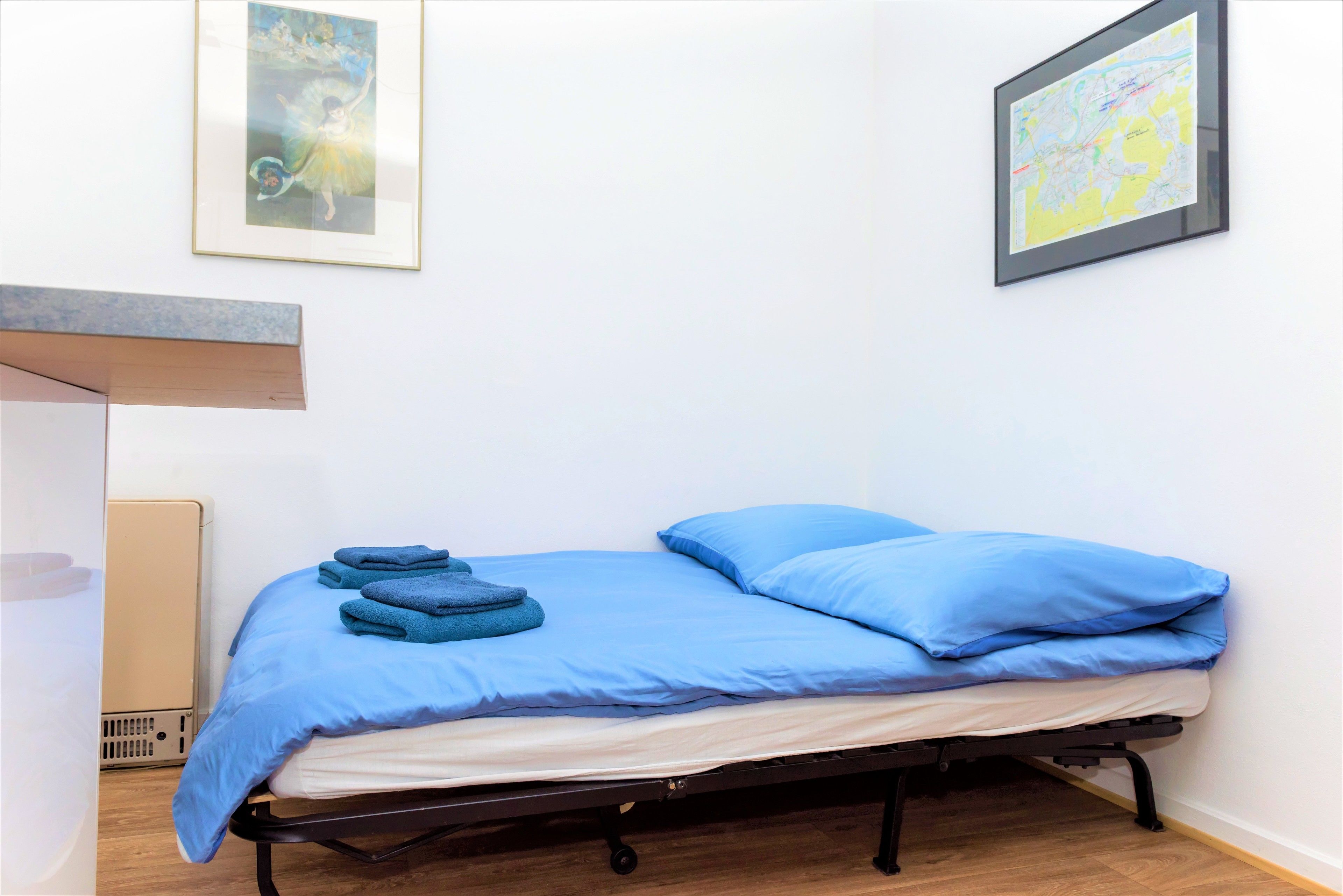 Un letto singolo con biancheria azzurra, un pavimento in legno e una parete bianca con due quadri appesi.