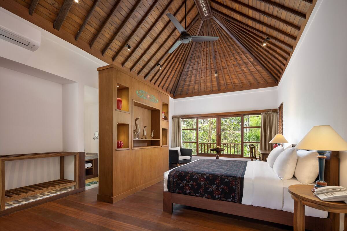 A spacious bedroom with a large bed, wooden ceiling beams, a ceiling fan, and a view of greenery through the window.