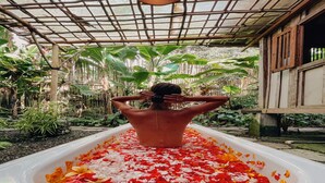 Eine Person in einer Badewanne, gefüllt mit Blütenblättern, umgeben von üppigem Grün und einer Bambuskonstruktion.