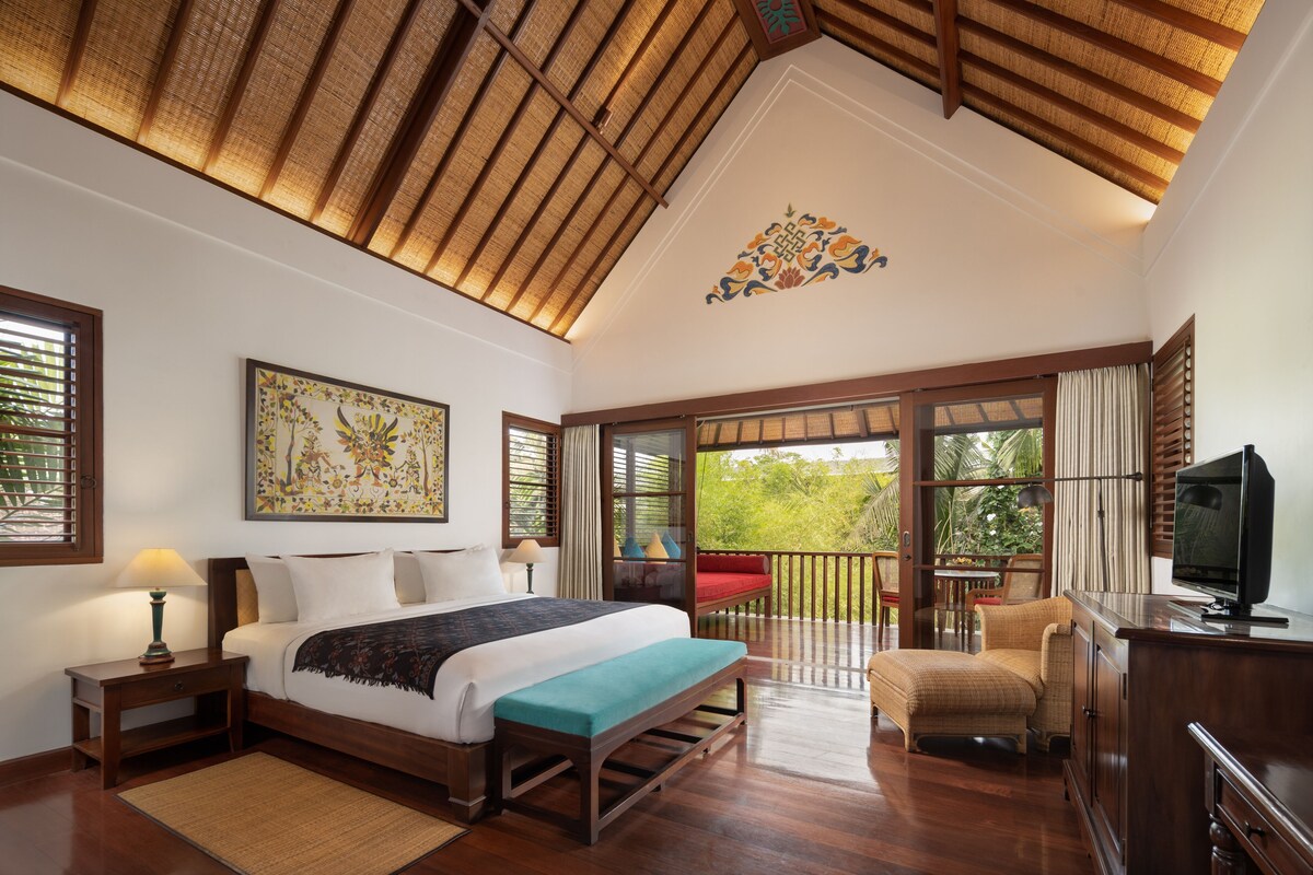 A spacious bedroom with a large bed, a bench, a television, and a balcony with a view of greenery.