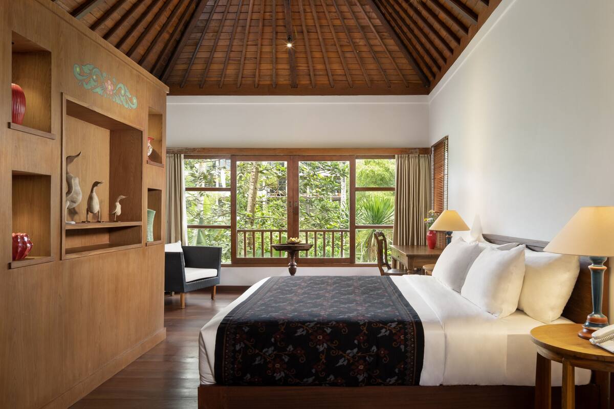 A spacious bedroom with a large bed, wooden shelves, a desk, and a balcony with a view of greenery.