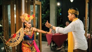 A person in traditional attire holding a fan, standing in an outdoor area with a table and chairs in the background. - Korurua Dijiwa Ubud (Ubud)