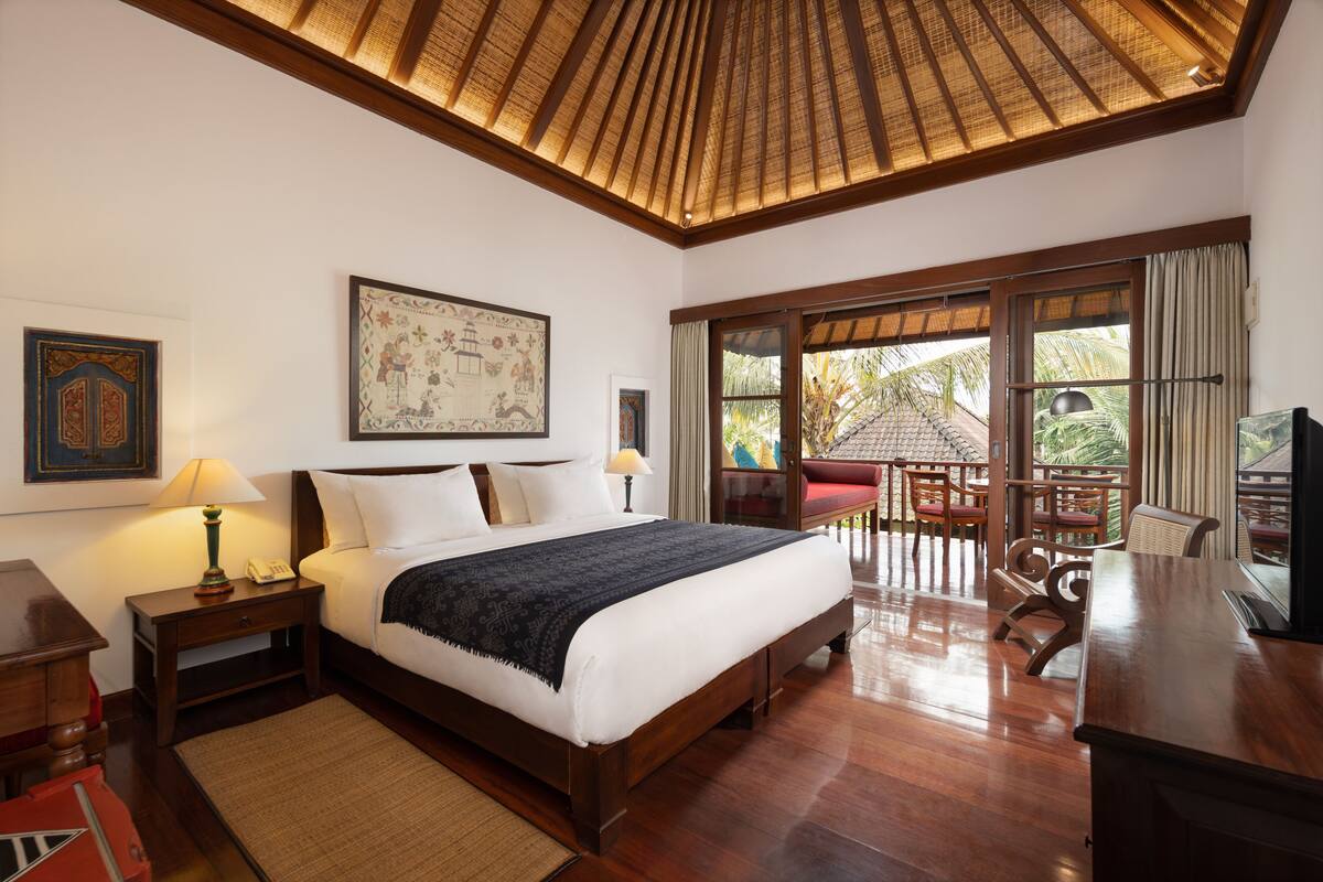 A spacious bedroom with a large bed, wooden headboard, and a ceiling with a woven design. There is a desk with a chair and a television, and a balcony with outdoor seating.