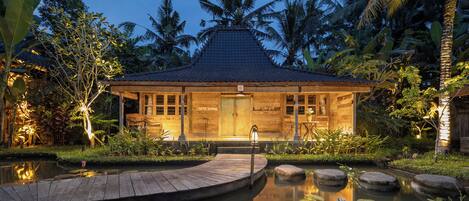 A wooden house with a tiled roof, surrounded by a pond with lily pads and a wooden dock.