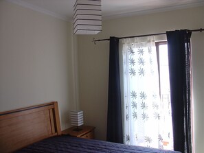 Quarto com cama, mesa de cabeceira, abajur e janela com cortinas.