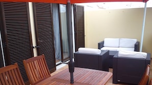 Eine Terrasse mit einem Holztisch und Stühlen, ein Rattansofa mit weißen Kissen und eine Glastür.