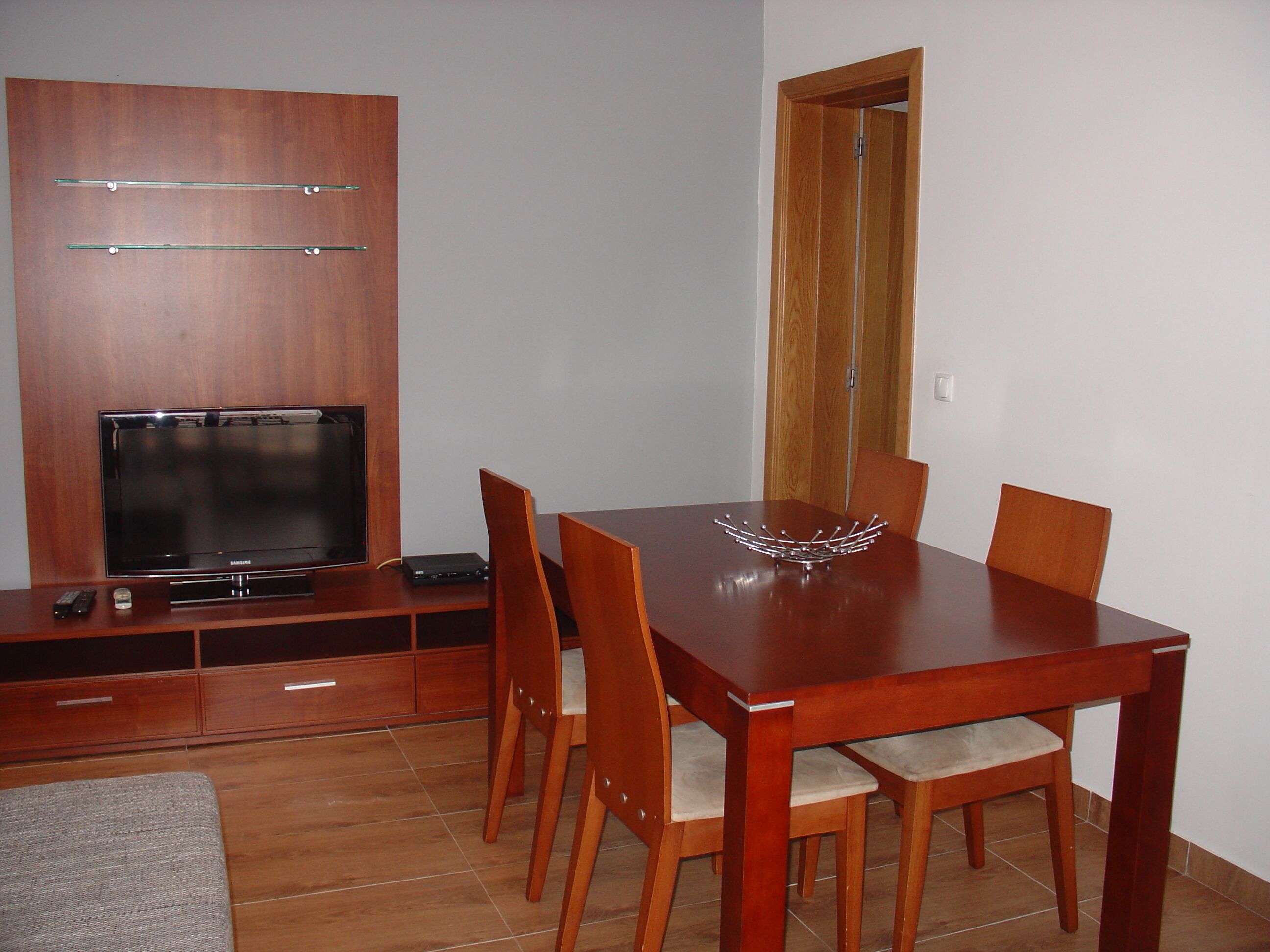 A dining area with a wooden table and chairs, a television on a wooden cabinet, and a door in the background.