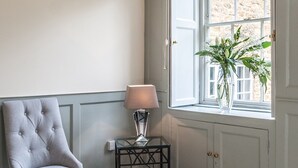 A corner room with a grey upholstered chair, a glass table with a lamp, and a window with a plant.