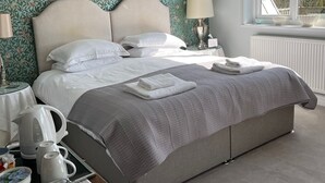 Una cama bien tendida con un edredón gris, una mesita de noche con una lámpara, un hervidor y un cuenco de fruta.