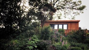 Una cabaña de madera con un frente con ventanas, rodeada de follaje denso y diversas plantas.