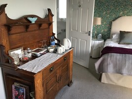 Una habitación con un aparador de madera, una cama con una colcha blanca y morada, una mesita de noche con una lámpara y una puerta con papel tapiz floral.