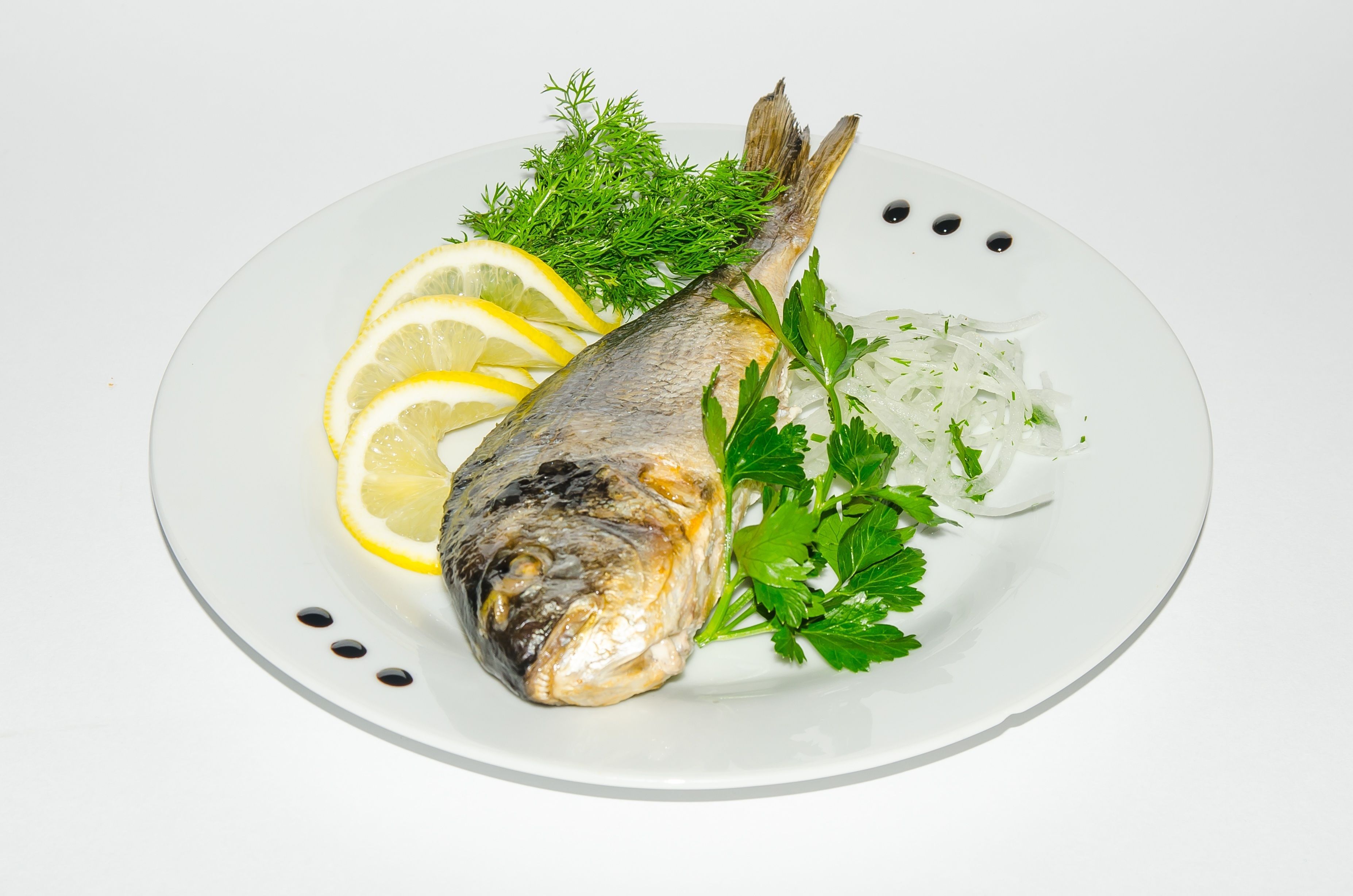 A plate with a whole fish, lemon slices, parsley, and dill.