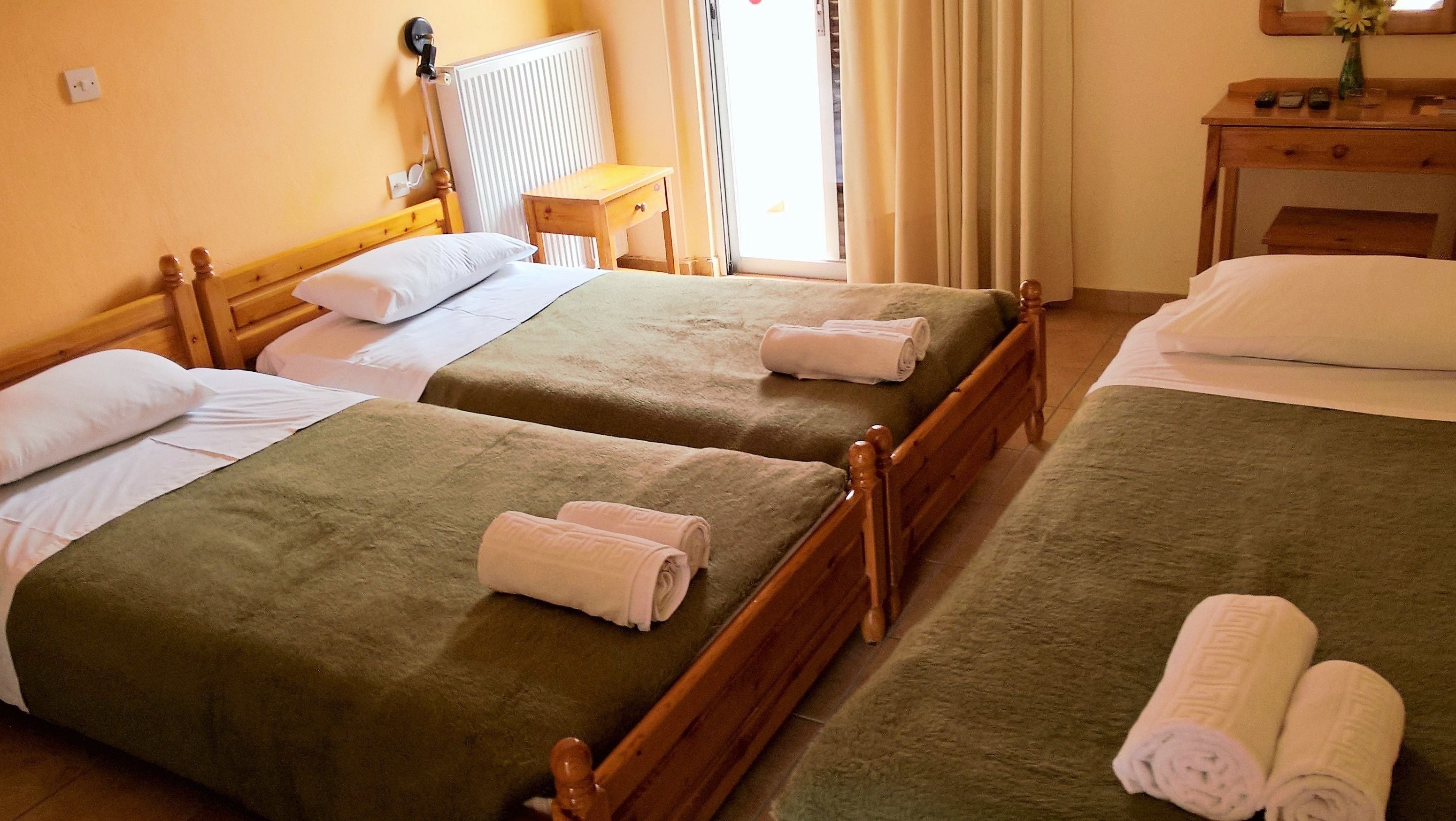 Een hotelkamer met twee eenpersoonsbedden, elk met een groene spreidek en witte kussens. Er staat een houten nachtkastje met een bloevas, een spiegel en enkele spullen erop.