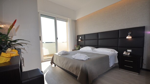 Une chambre d’hôtel moderne avec un grand lit, une table de chevet avec une lampe, une petite commode et une vue sur le balcon.