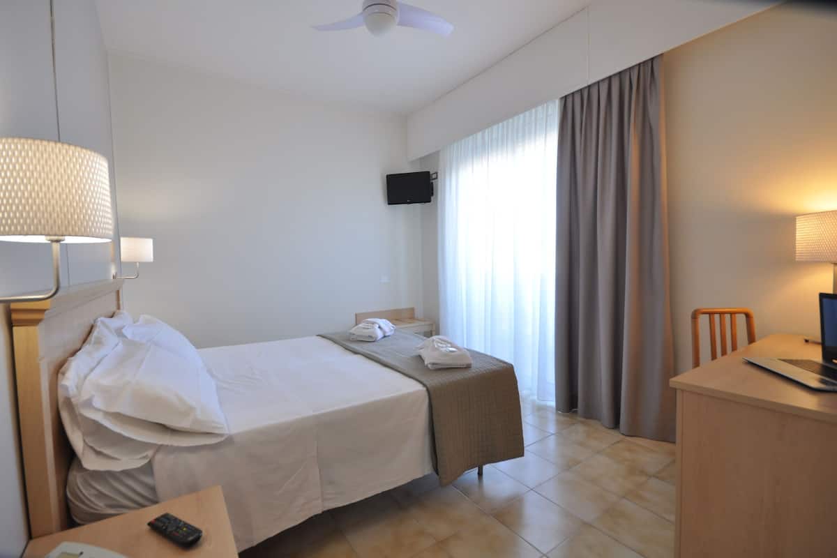 A hotel room with a bed, a desk with a laptop, a TV on the wall, and a ceiling fan.