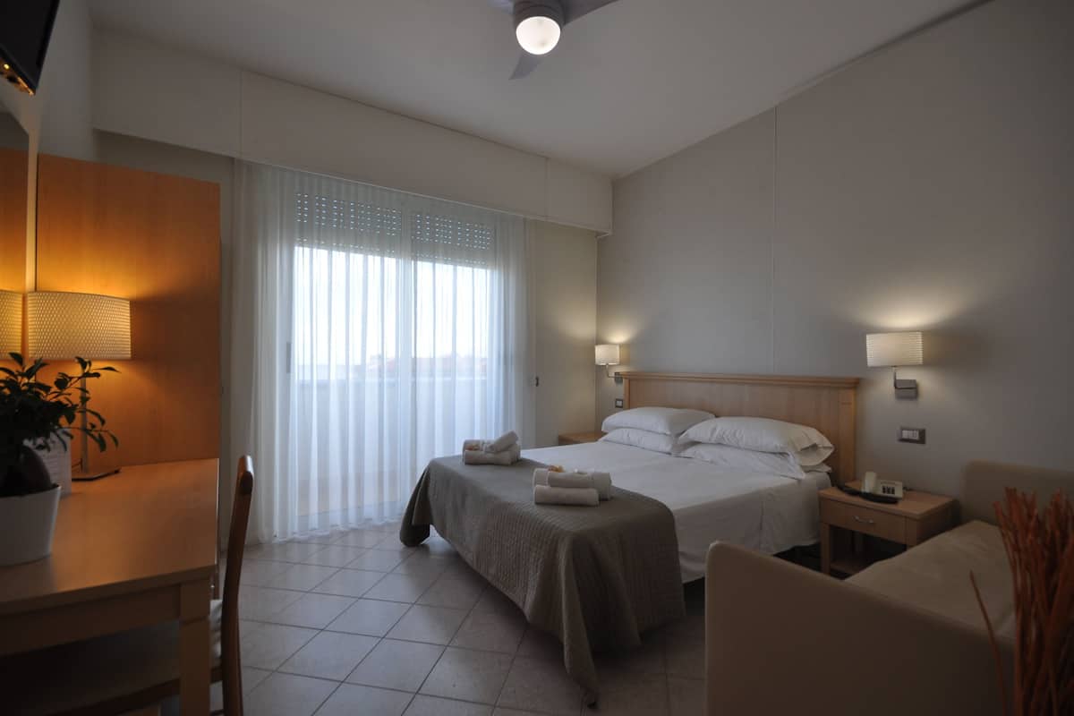 A hotel room with two beds, a desk, a chair, a lamp, and a window with blinds.