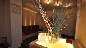 Un espace d’accueil avec un vase en verre transparent contenant des branches et des fleurs rouges, posé sur une table éclairée, une horloge au mur et un extincteur sur le sol.
