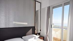 Une chambre à coucher comprenant un lit, une table de chevet, un miroir et une porte-fenêtre coulissante donnant sur un balcon avec vue sur la mer.