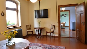 Quarto com uma mesa redonda de madeira, duas cadeiras, um vaso com flores, uma televisão de tela plana na parede e uma porta de madeira que dá para outro cômodo.