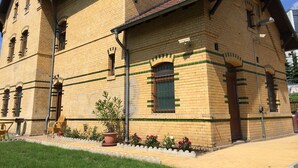 Um edifício de dois andares com fachada de tijolos, várias janelas e uma pequena área de jardim com vasos de plantas e flores.