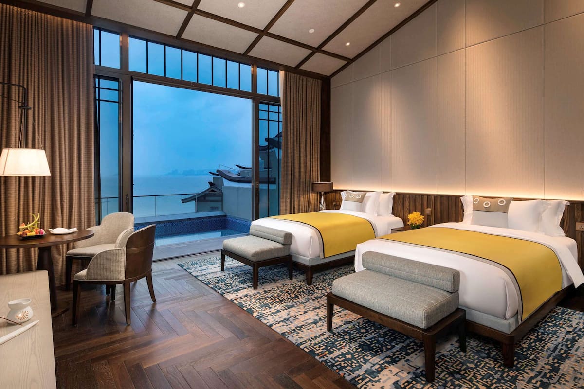 Une chambre d’hôtel avec deux lits, un bureau et une chaise. De grandes fenêtres offrent une vue sur la mer.