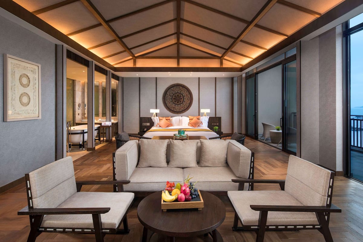 Une chambre d’hôtel moderne avec un grand lit, deux canapés, une table basse avec des fruits et une vue sur la mer.