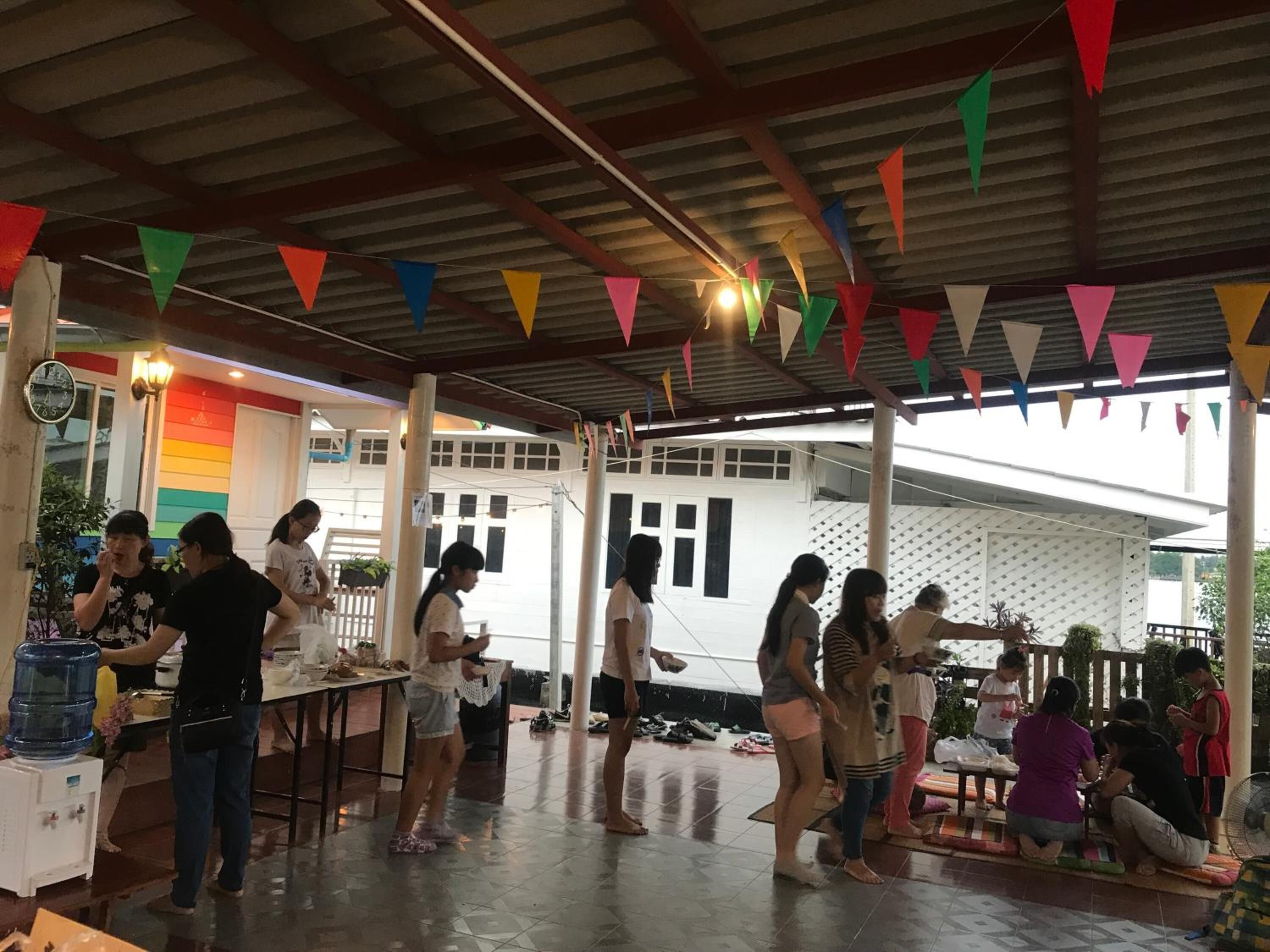 Un espacio al aire libre techado con banderas triangulares de colores, un dispensador de agua y gente reunida alrededor de mesas.