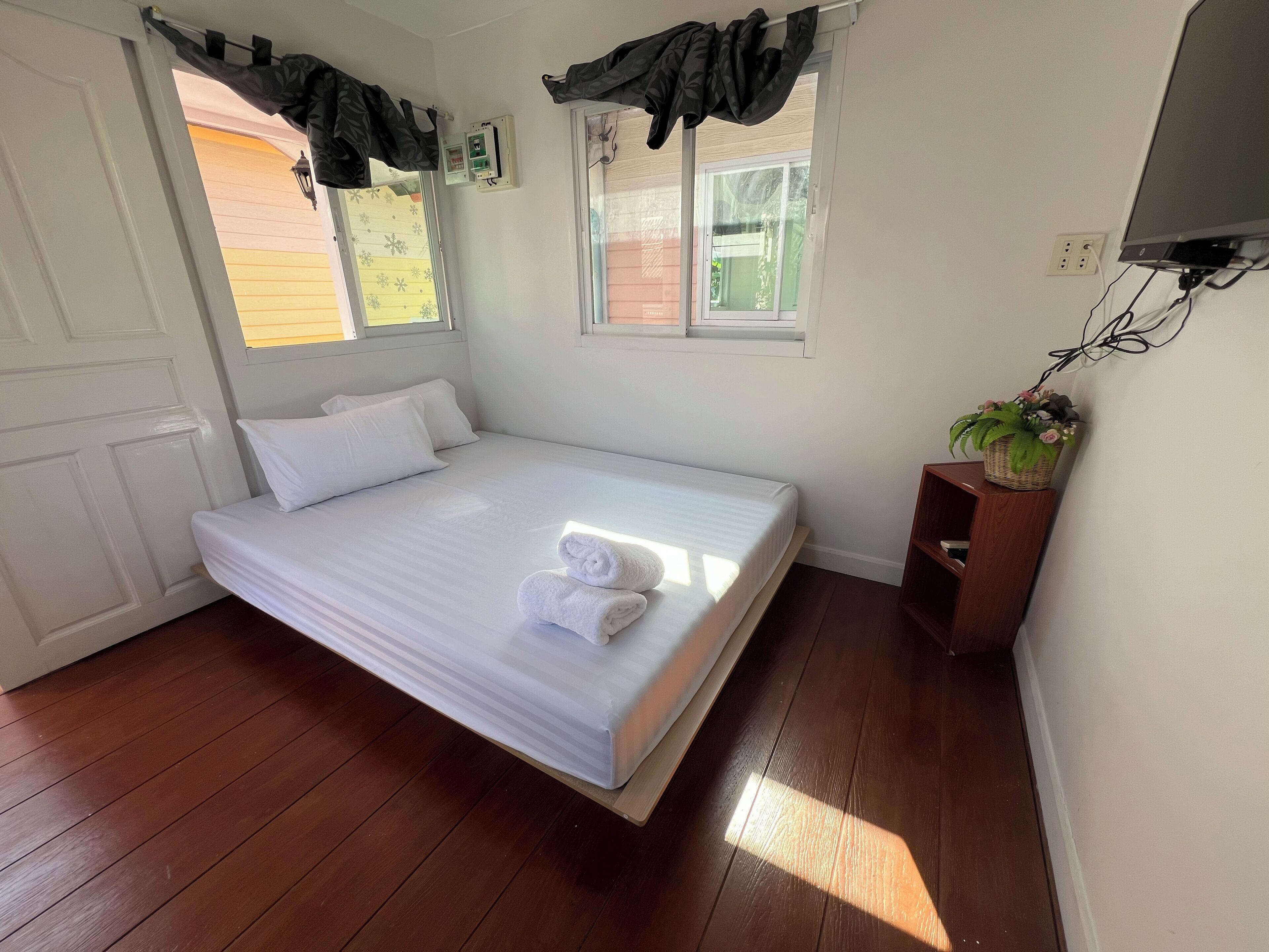 A single bed with white linens, a wooden floor, a window with curtains, a wall-mounted TV, and a small wooden shelf with a plant.