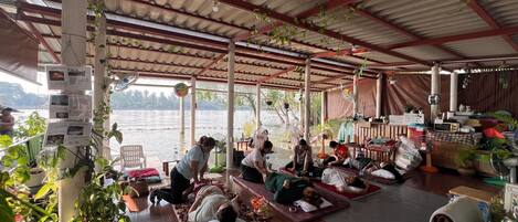 A covered outdoor seating area with people resting on mats, surrounded by potted plants and various items.