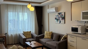 A modern living room with a sofa, coffee table, and a dining area with a table and chairs. There is a kitchen area with a microwave and cabinets. - Upart Home (Mersin)