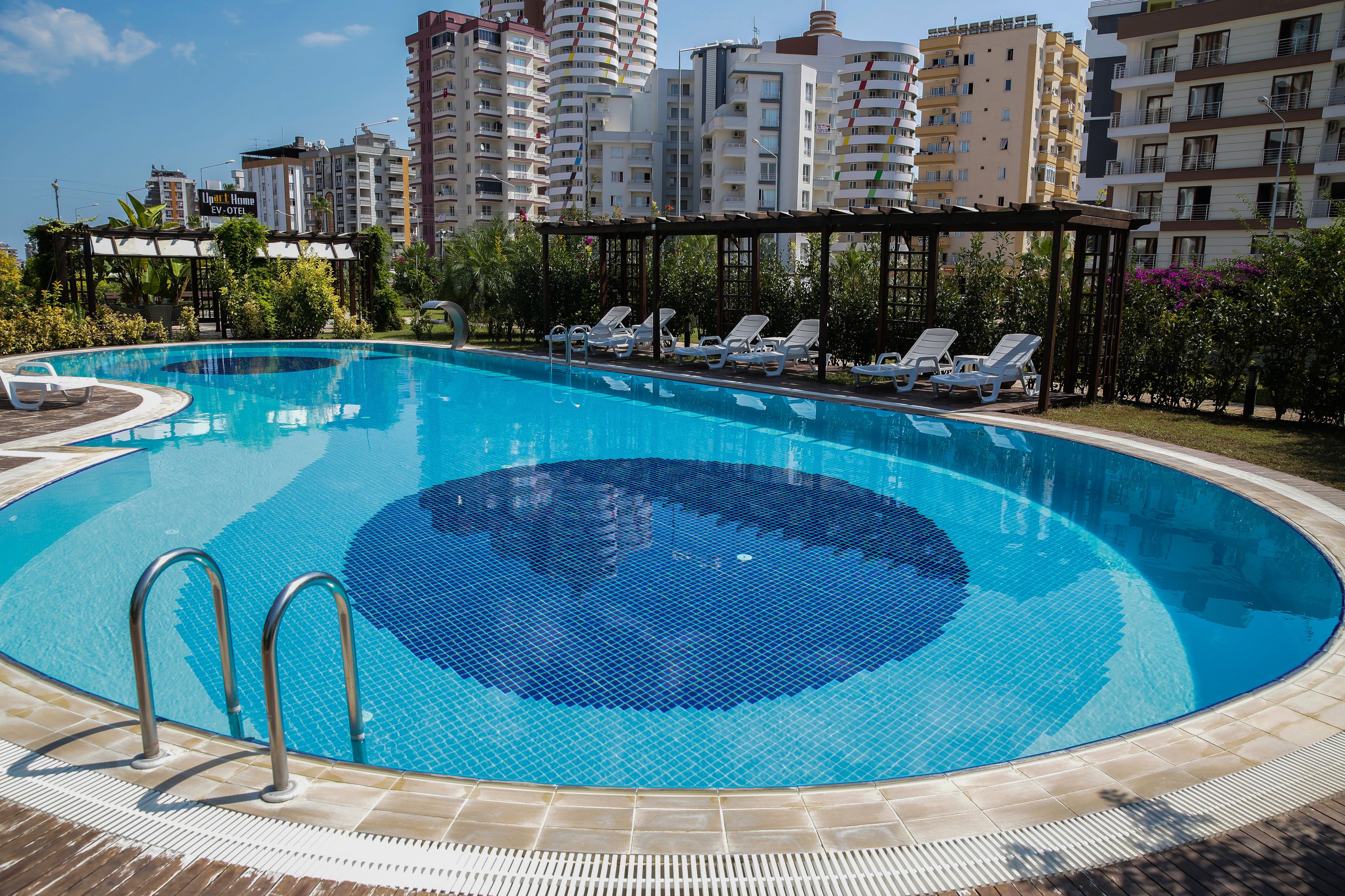 A swimming pool with lounge chairs and a pergola, surrounded by greenery and tall buildings in the background.