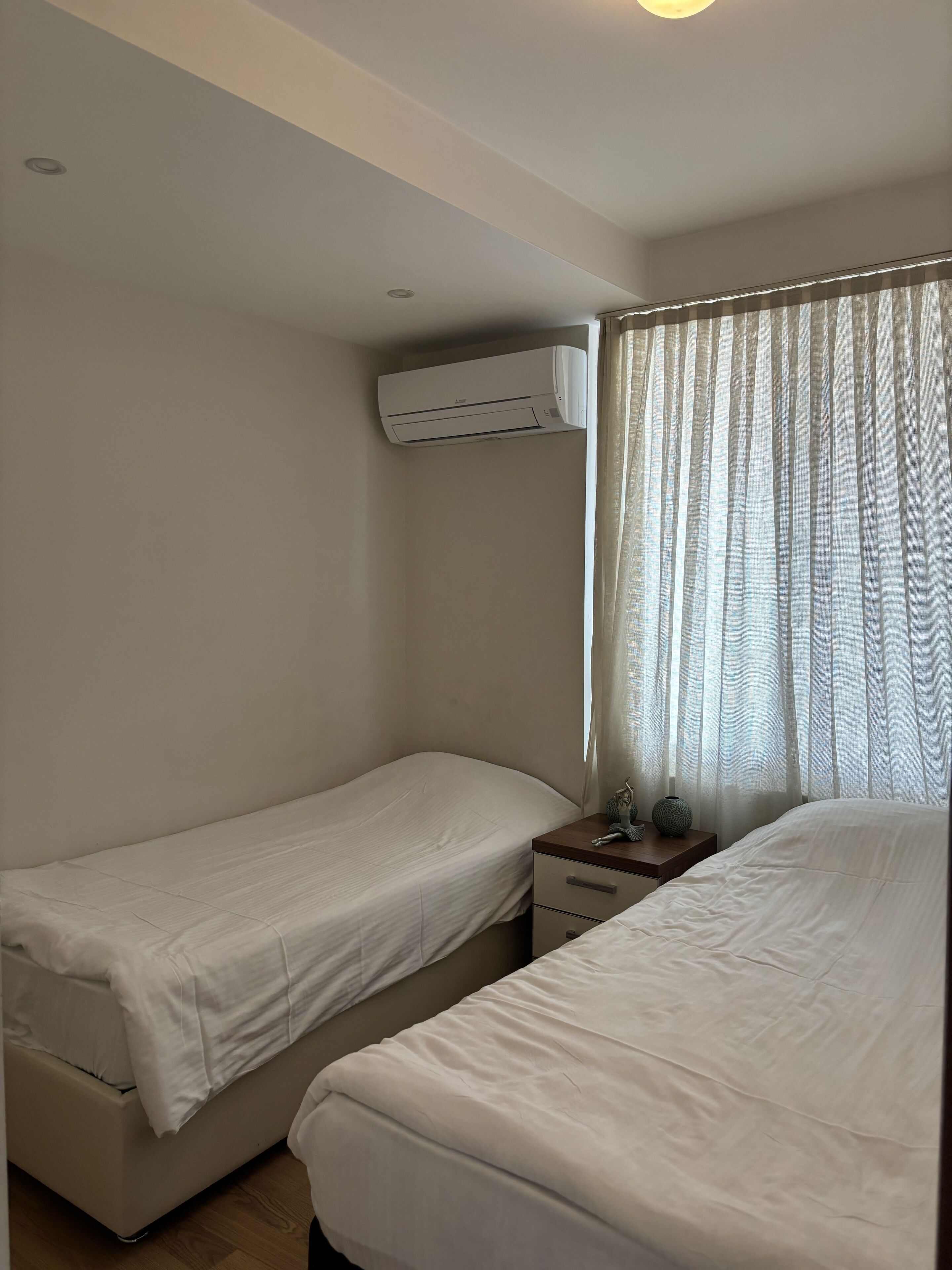Una habitación con dos camas individuales, una mesita de noche con un jarrón y una pequeña escultura, y un equipo de aire acondicionado instalado en la pared.