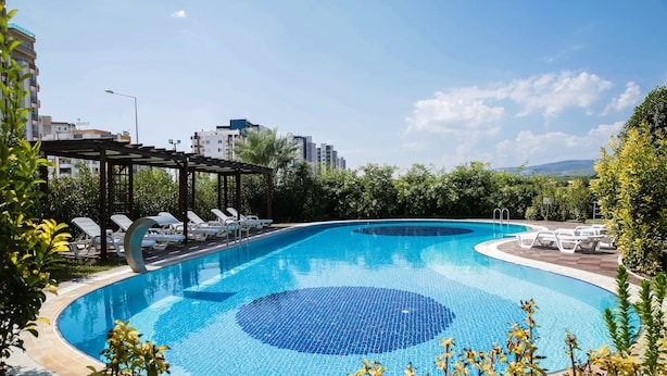 A swimming pool with lounge chairs and a pergola, surrounded by greenery and buildings in the background. - Upart Home (Mersin)