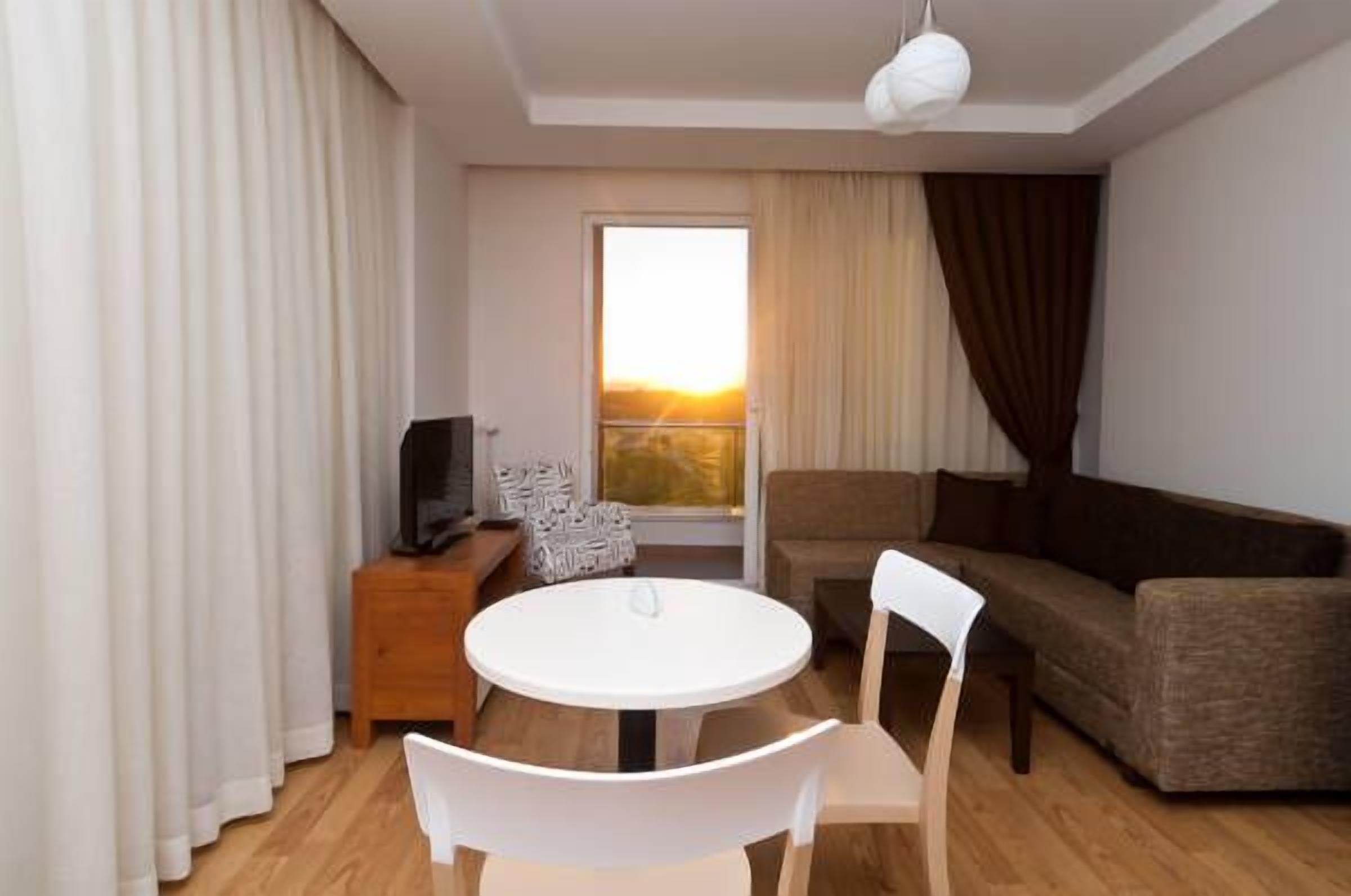 Un salón moderno con una mesa redonda blanca, dos sillas blancas, un sofá marrón, un televisor sobre un mueble de madera y un amplio ventanal con cortinas.