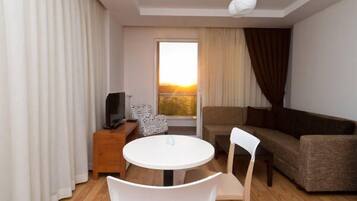 Un salón moderno con una mesa redonda blanca, dos sillas blancas, un sofá marrón, un televisor sobre un mueble de madera y un amplio ventanal con cortinas.