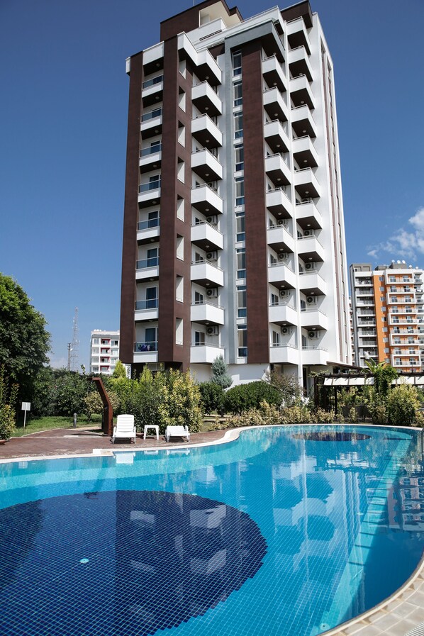 A swimming pool with a tiled deck, surrounded by greenery and a tall residential building in the background. - Upart Home (Mersin)