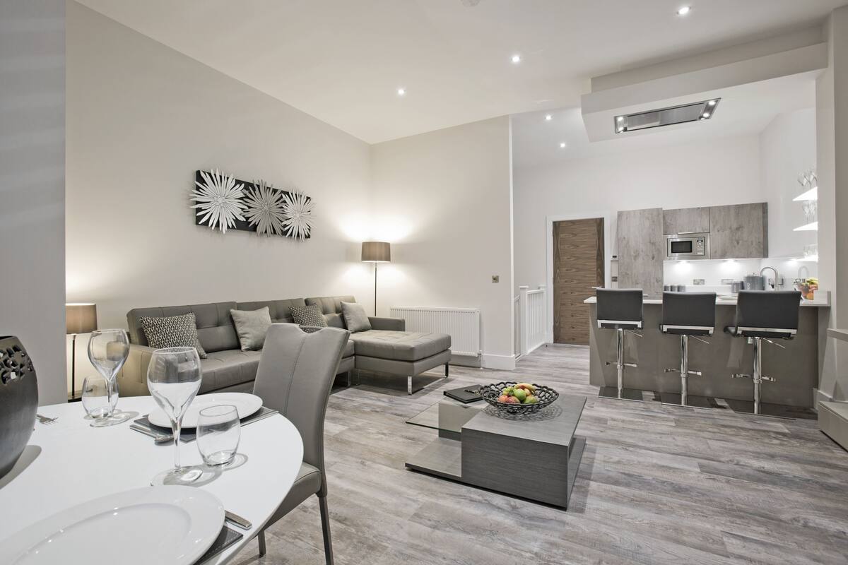 Un salon moderne comprenant un coin repas, des canapés gris, une table basse et une cuisine avec des tabourets de bar.