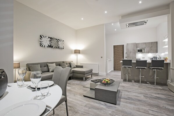 A modern living room with a dining area, grey sofas, a coffee table, and a kitchen with bar stools. - Apple Apartments Devanha Gardens (Aberdeen)